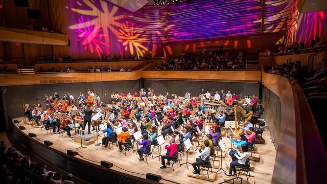 The photo captures a vibrant orchestral performance in progress inside a modern concert hall. The musicians are seated in a semi-circular arrangement on stage, surrounded by wood-paneled walls that reflect the colorful, abstract light patterns projected above them, creating a dynamic and festive atmosphere. A conductor is visible at the forefront, leading the orchestra. The audience, seated in the curved balconies and the main floor, is attentively watching the performance in a well-lit, packed venue.
